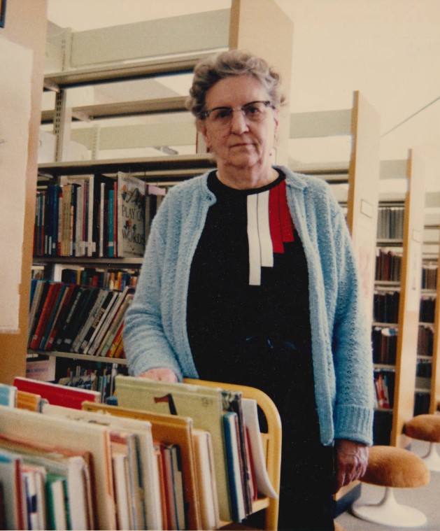 Marian Parker in front of a stack of books, wearing a blue cardigan and glasses.